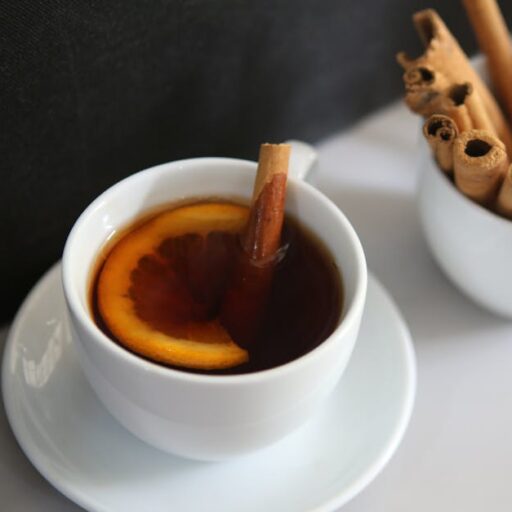 A white cup of cinnamon-flavored tea with a citrus slice, accompanied by cinnamon sticks.