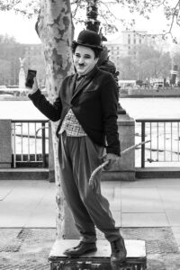 Black and white photo of a street performer dressed as Charlie Chaplin on a London riverside.